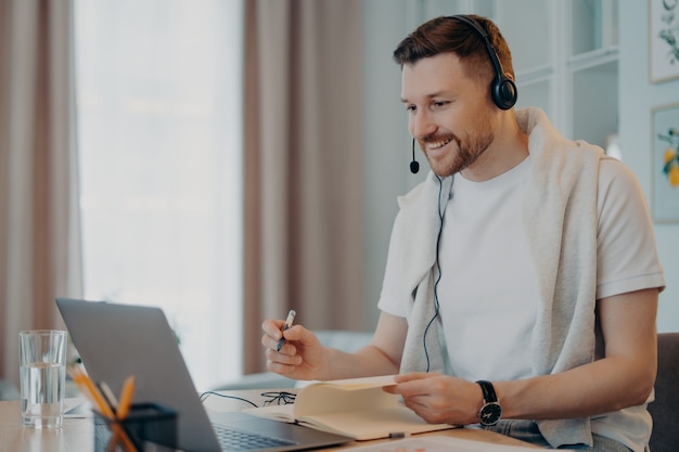 Ainda bem que o tutor masculino dá aulas online e se comunica à distância com o trainee usa fones de ouvido com microfone e o laptop faz anotações necessárias em poses de diário no espaço de coworking. e processo de aprendizagem