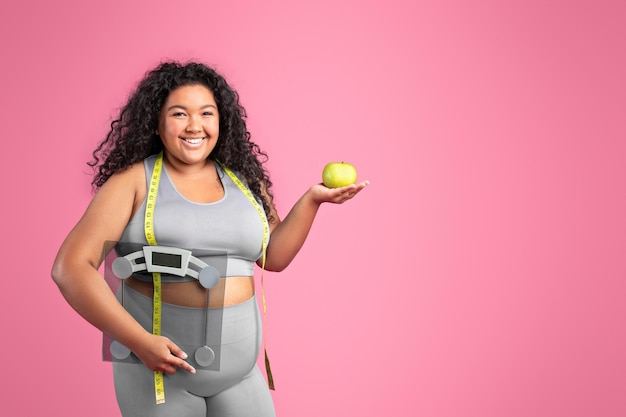Foto ainda bem que uma mulher negra positiva em roupas esportivas segurando maçã e balança aproveitando o resultado da dieta rosa