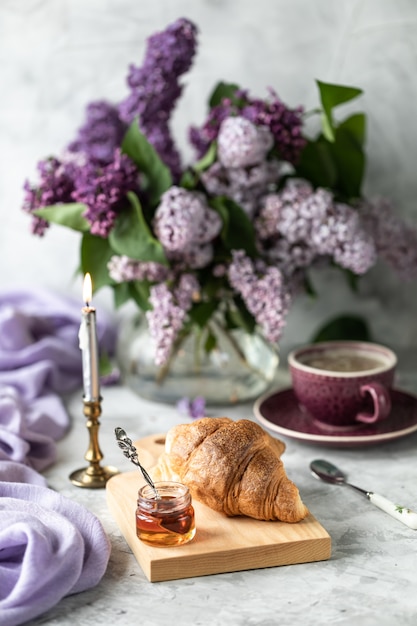 Ainda croissants de vida, uma xícara de café e um buquê de lilases na mesa perto da janela.