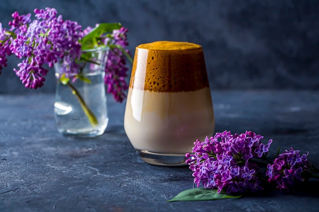 Ainda vida de verão com café dalgona em copo alto e flores lilás no escuro. Café instantâneo batido com açúcar e água e adicionado ao leite frio.