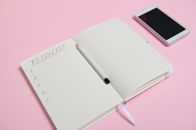 Ainda vida. Um telefone celular branco deitado sobre um fundo rosa ao lado de um lápis no meio de uma agenda aberta, diário, caderno com uma lista para fazer em uma folha de papel branca alinhada com o espaço da cópia