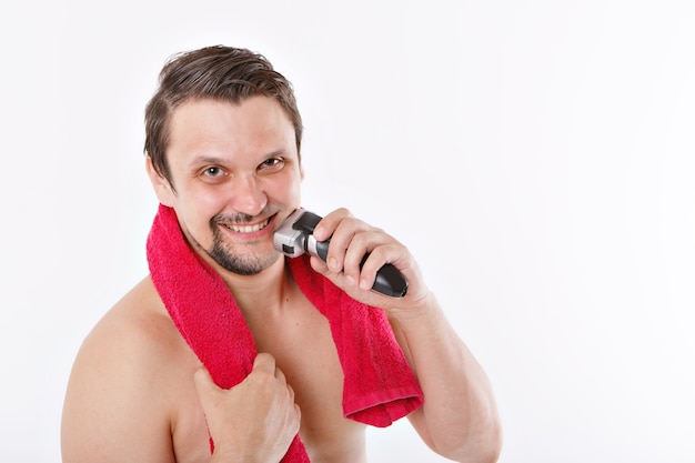 Aislado: un hombre se afeita la barba. el tipo se limpia la barba con una maquinilla de afeitar eléctrica. tratamientos matutinos en el baño. toalla roja alrededor de su cuello. copia espacio