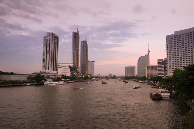 Ajardine la vista de la puesta del sol en el río Chao Phraya con una vista de barcos y de edificios modernos a lo largo de la orilla.