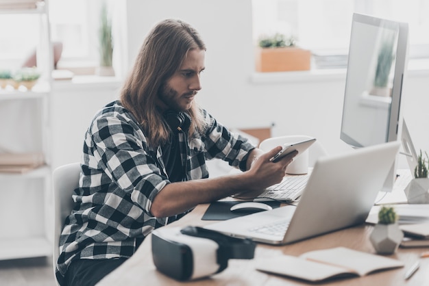 Ajetreado día de trabajo. Apuesto joven con cabello largo trabajando en la tableta digital mientras está sentado en su escritorio en la oficina creativa