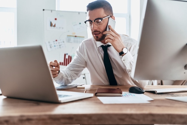 Ajetreado día de trabajo. Apuesto joven en ropa formal hablando por teléfono mientras está sentado en la oficina