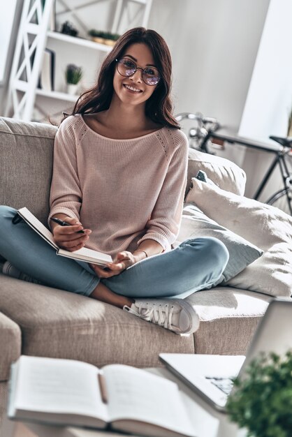 Ajetreado día de trabajo. Hermosa mujer joven en gafas estudiando y sonriendo mientras está sentado en el sofá en casa