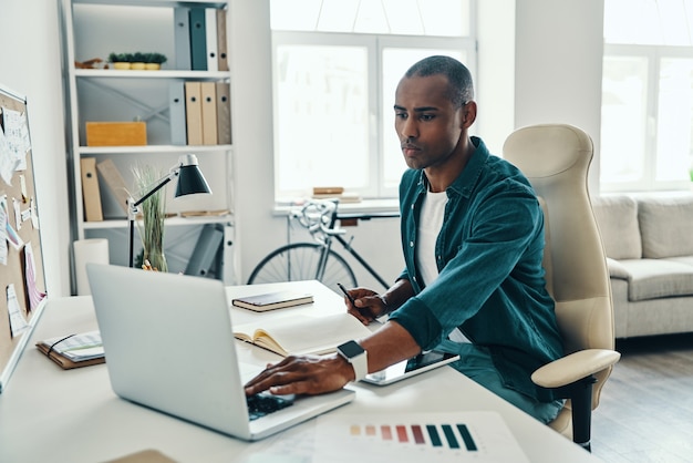 Ajetreado día de trabajo. Hombre africano joven pensativo en camisa usando la computadora portátil mientras está sentado en la oficina