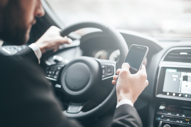 Ajetreado día de trabajo. Primer plano de joven empresario escribiendo algo en el teléfono inteligente mientras conduce un coche