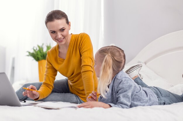 Ajude-me. Mãe otimista e vigorosa trabalhando em um laptop enquanto sorri e fala com a filha