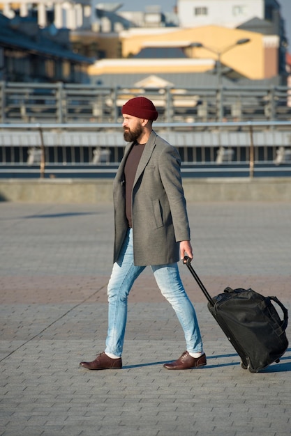 Ajustar la vida en la nueva ciudad Hombre barbudo hipster viajar con bolsa de equipaje sobre ruedas Viajero con maleta llegar aeropuerto estación de tren fondo urbano Hipster listo disfrutar viajar Llevar bolsa de viaje