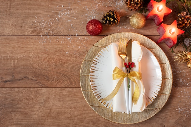 Ajuste de lugar de jantar de férias de Natal - pratos, guardanapo, talheres, decorações de ouro bugiganga sobre a mesa de carvalho. Garfo e colher em placas de ouro. Em torno de velas vermelhas, cones e bolas. Configuração plana