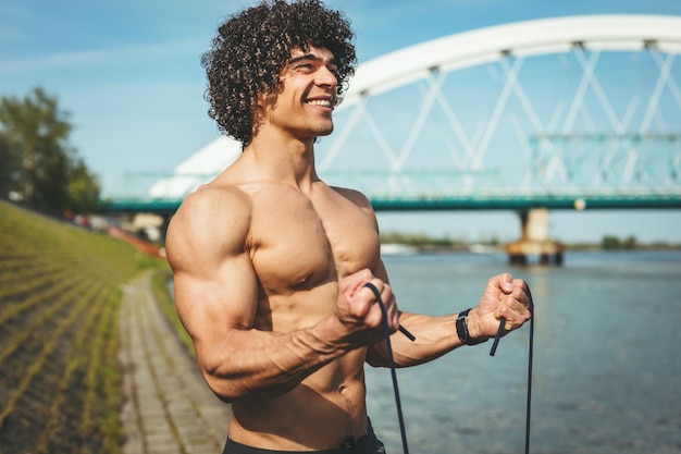 Ajuste jovem musculoso com torso nu fazendo exercício de bíceps com banda de resistência perto do rio.