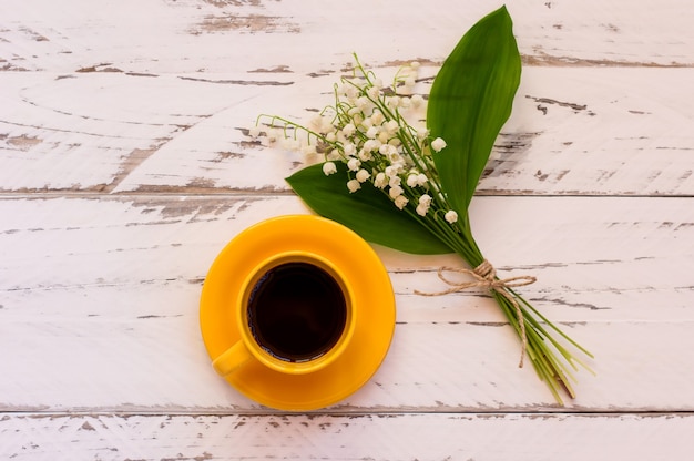 Ajuste de la mesa de café de la mañana con ramo de flores de lirio de los valles. Taza amarilla con café negro decorado con flores de primavera en la mesa de madera, vista superior.