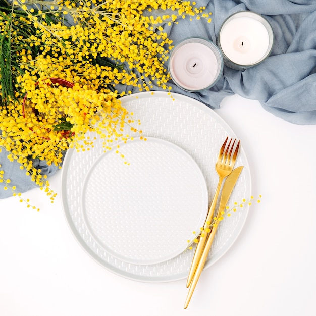 Ajuste de la mesa festiva. Platos y cubiertos con textil decorativo gris y flores amarillas sobre fondo blanco. Hermoso arreglo plano.