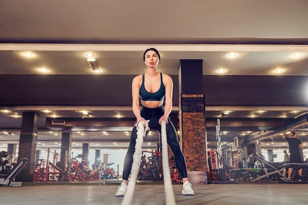 Ajuste mujer vestida con ropa deportiva posando con cuerdas de batalla en el gimnasio