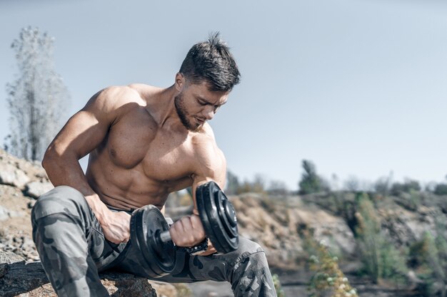 Ajuste o jovem levantando halteres, fazendo exercícios ao ar livre. Esporte, fitness, levantamento de peso, musculação, treinamento, atleta, conceito de exercícios de treino. Vista lateral.