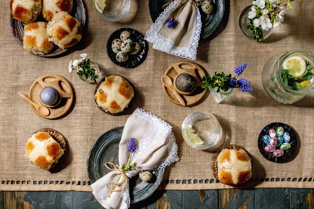 Ajuste de la tabla de Pascua con huevos de colores y chocolate, bollos cruzados calientes, ramo de flores, plato de cerámica vacío con servilleta, vaso de bebida de limonada en la mesa de madera con mantel textil