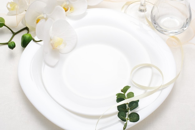 Ajuste de la tabla romántica de vacaciones con orquídeas, plato vacío blanco y cinta de seda sobre fondo blanco, ajuste de la mesa de boda