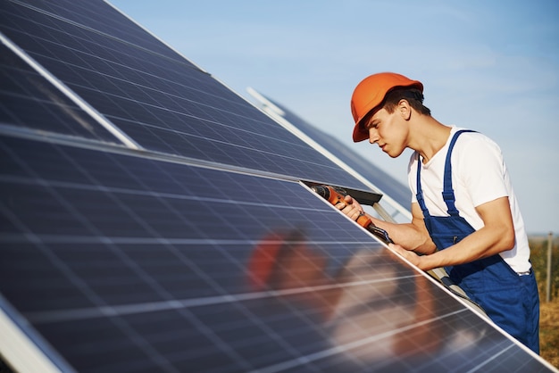 Akkuschrauber verwenden. Männlicher Arbeiter in blauer Uniform im Freien mit Solarbatterien am sonnigen Tag.