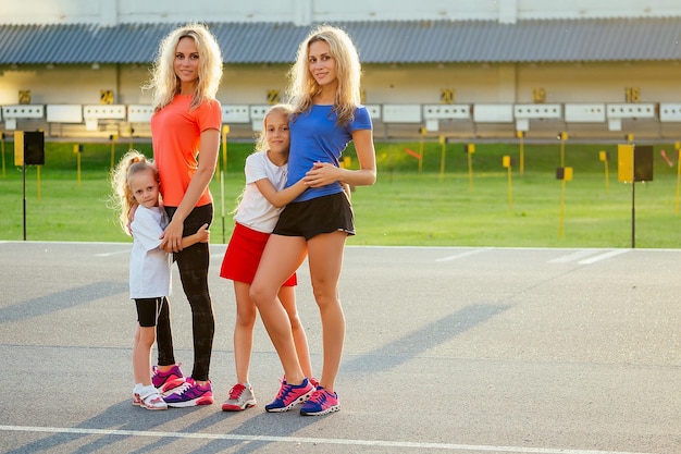 Aktive Familie im Freien. zwei blonde zwillingsschwestern und ihre töchter in stylischer sportbekleidung wärmen sich auf und machen übungen crossfit und fitness im stadion. Gruppe von vier Mädchen im Park.