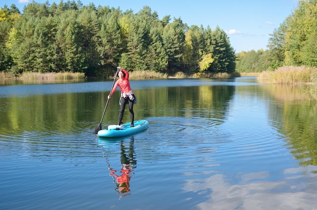 Aktive Frau, die SUP-Brett auf schönem See, Herbstwaldlandschaft und Natur im Hintergrund paddelt