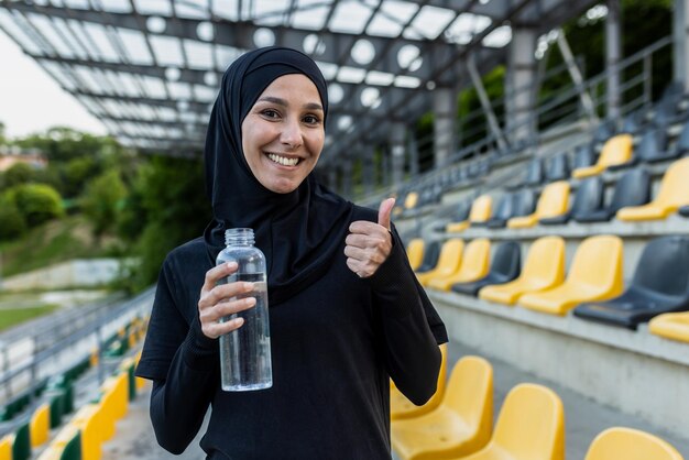 Aktive Frau im Hijab, die eine Wasserflasche in der Hand hält und einen Daumen nach oben in einem Außenstadion zeigt