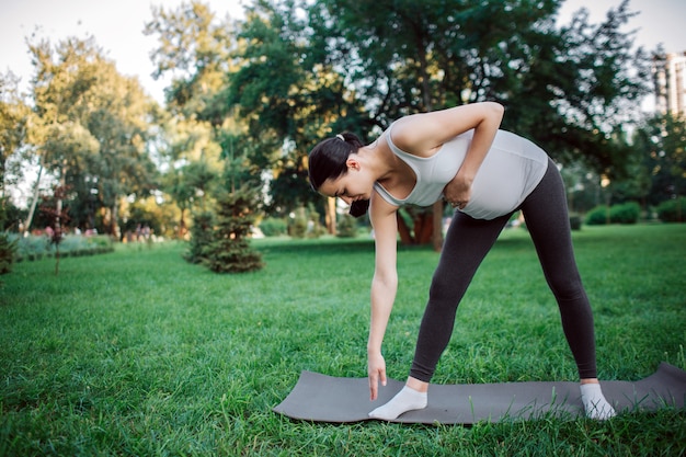 Aktive junge schwangere Frau, die auf Yogakameraden im Park trainiert. Sie erreicht den Boden mit einer Hand und hält eine andere am Bauch.