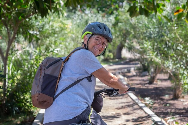 Aktive lächelnde Seniorin, die mit ihrem Elektrofahrrad in einem grünen Park mit Helm im Rückblick radelt Reife attraktive Frau, die den Ruhestand und einen gesunden Lebensstil genießt Tropischer Park