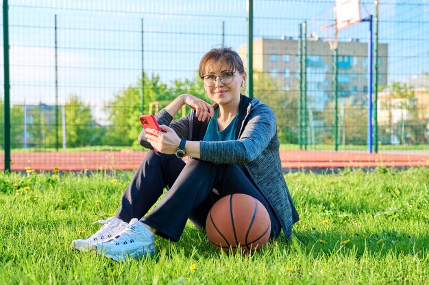 Aktive reife Frau mit Basketball und Smartphone im Freien Basketballplatz Hintergrund Frau mittleren Alters in Sportbekleidung Kopfhörer sitzt auf dem Gras Sportliche gesunde Lebensweise Freizeitaktivität