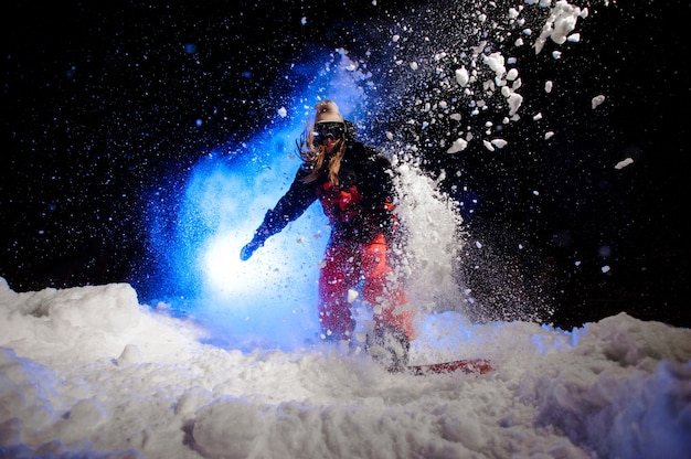 Aktive Snowboarderin in roter Sportbekleidung, die nachts unter blauem Licht auf dem Berghang im Schnee springt