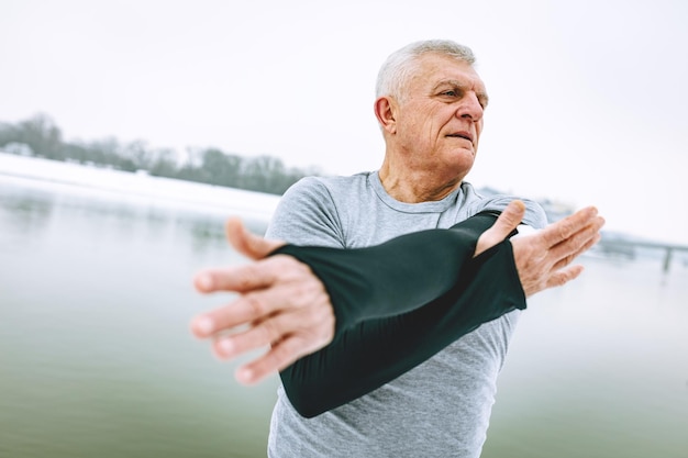 Aktiver älterer Mann, der sich während des Wintertrainings im Freien am Fluss dehnt und Übungen macht.