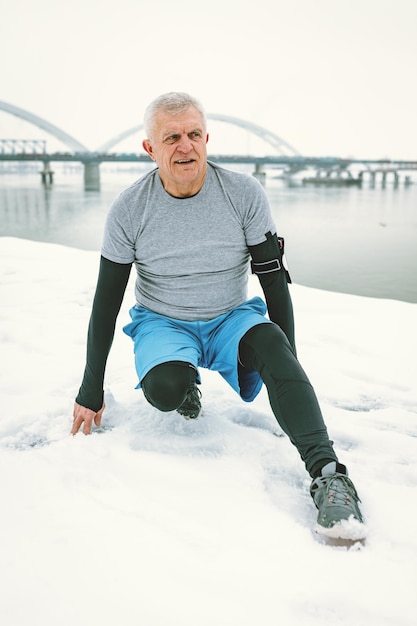 Aktiver älterer Mann, der sich während des Wintertrainings im Freien am Fluss dehnt und Übungen macht.