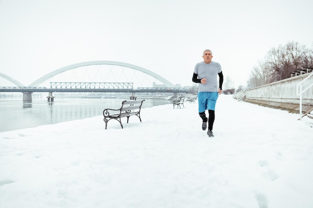 Aktiver älterer Mann, der während des Wintertrainings draußen am Fluss läuft und Übungen macht. Platz kopieren.
