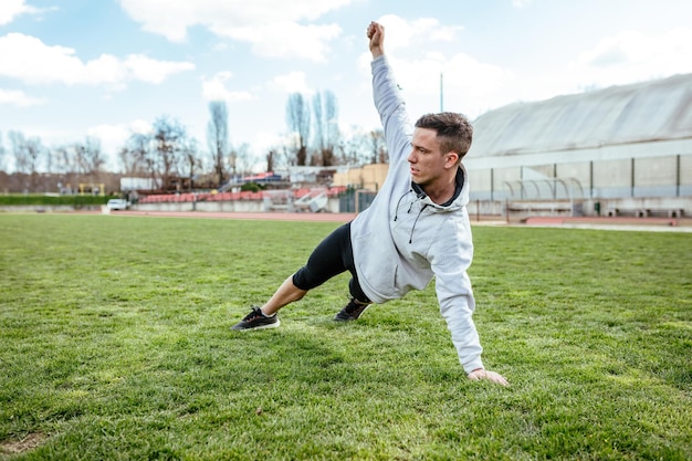 Aktiver junger Mann, der sich im leeren Stadion auf dem öffentlichen Platz dehnt und Übungen macht.