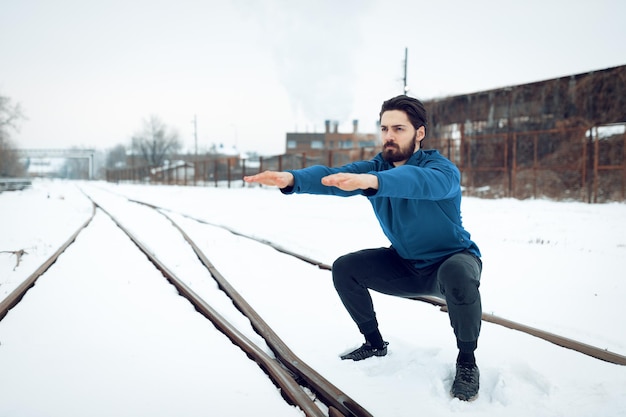 Aktiver junger Mann, der während des Wintertrainings draußen in der Öffentlichkeit unter alten Eisenbahnen hockt und Übungen macht. Platz kopieren.