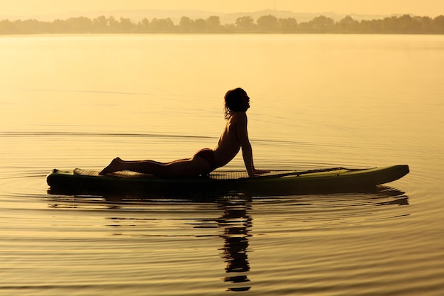 Aktiver junger Mann in Silhouette, der den Körper auf dem Sup-Board dehnt