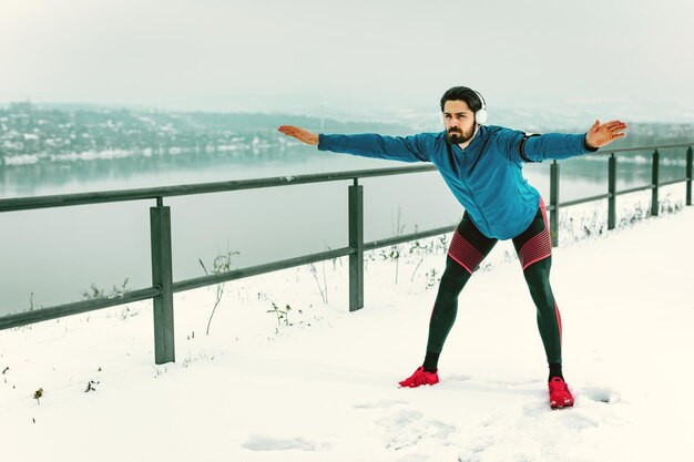 Aktiver junger Mann mit Kopfhörern an den Ohren, der sich während des Wintertrainings draußen an öffentlichen Orten dehnt und Übungen macht.