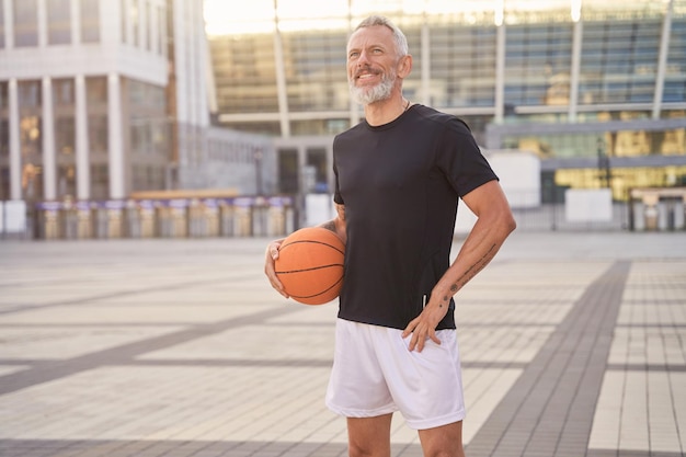 Aktiver Mann mittleren Alters in Sportkleidung, der mit Basketballball im Freien steht, der bereit ist für