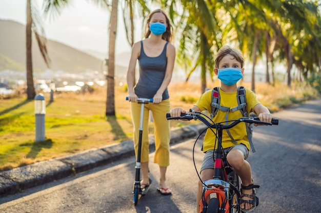 Aktiver Schuljunge und seine Mutter mit medizinischer Maske fahren an einem sonnigen Tag Fahrrad mit Rucksack. Fröhliches Kind radelt auf dem Weg zur Schule. Wegen der Coronavirus-Epidemie müssen Sie mit einer Maske zur Schule gehen