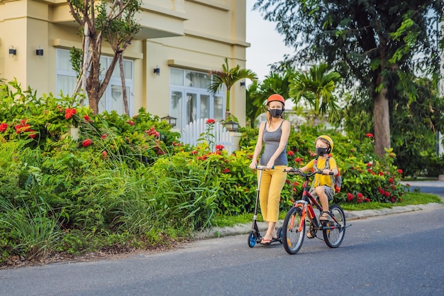 Aktiver Schuljunge und seine Mutter mit medizinischer Maske und Schutzhelm fahren Fahrrad mit Rucksack