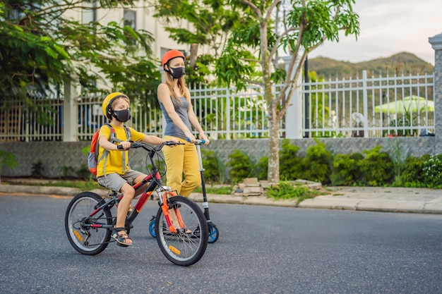 Aktiver Schuljunge und seine Mutter mit medizinischer Maske und Schutzhelm fahren Fahrrad mit Rucksack