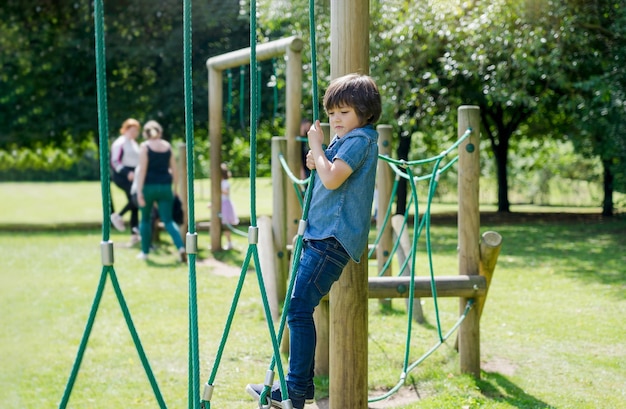 Aktives Kind hält Bademantel auf dem Spielplatz. Kind genießt Outdoor-Aktivitäten in einem Kletter-Abenteuerpark an einem sonnigen Sommertag. Niedlicher kleiner Junge, der Spaß auf einem öffentlichen Spielplatz hat