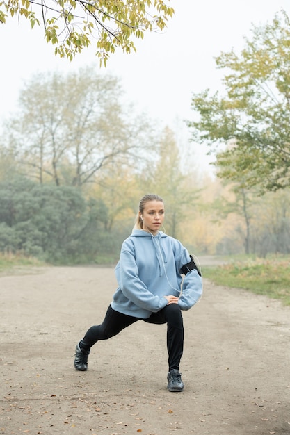 Aktives Mädchen in Kopfhörern, blauem Hoodie, schwarzer Leggins und Turnschuhen, die morgens Übungen zum Strecken der Beine auf dem Waldweg gegen Bäume machen