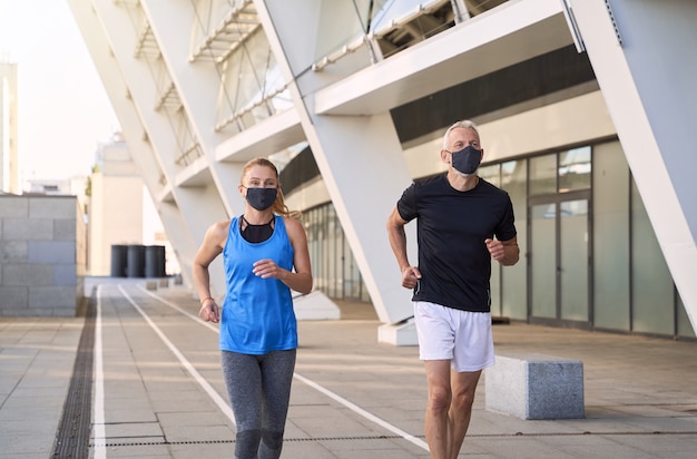 Aktives reifes Paar mit schützenden Gesichtsmasken, die zusammen in der städtischen Umgebung joggen