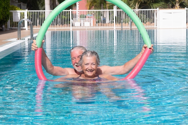 Aktives Seniorenpaar, das im Schwimmbad mit Schwimmnudeln Sport treibt Glückliche Rentner spielen im Außenpoolwasser unter der Sonne