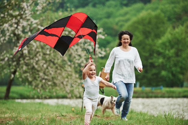 Aktives Spiel. Positives weibliches Kind und Großmutter, die mit rotem und schwarzem Drachen in den Händen draußen laufen