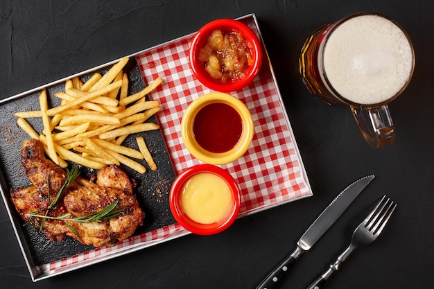 Alas de pollo al horno con papas fritas en una bandeja de hierro con vaso de cerveza sobre fondo negro. Naturaleza muerta. Foto de la carta del restaurante