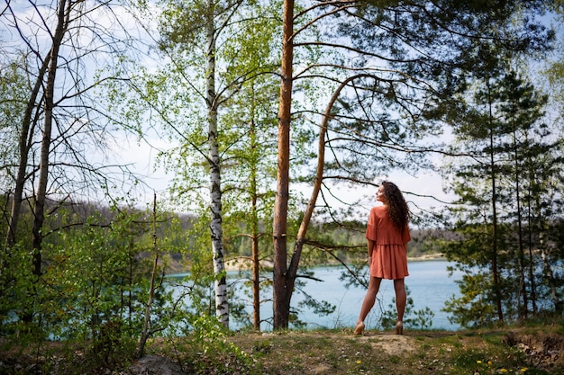 Albañilería de madera. Una niña con el pelo largo y ondulado y rizado con un vestido de guipur naranja y zapatos en la naturaleza, en un bosque junto al lago, estaba parada cerca de árboles y arbustos. Mujer joven sonríe y disfruta de la vida