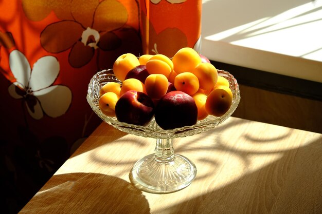 Albaricoques y melocotones maduros y jugosos en un frutero de vidrio en la mesa de madera cerca de la ventana soleada