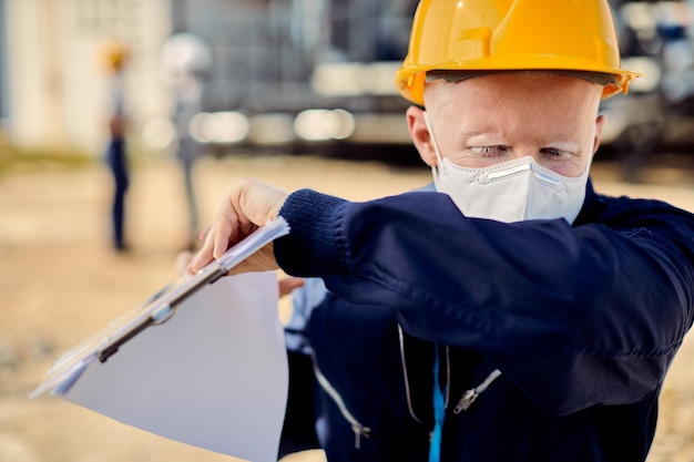 Albino-Bauingenieur mit Gesichtsmaske, der auf der Baustelle in den Ellbogen hustet